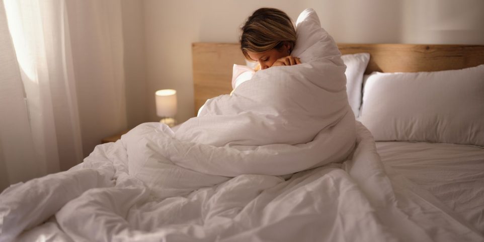 Depressed woman covered with duvet in bedroom.